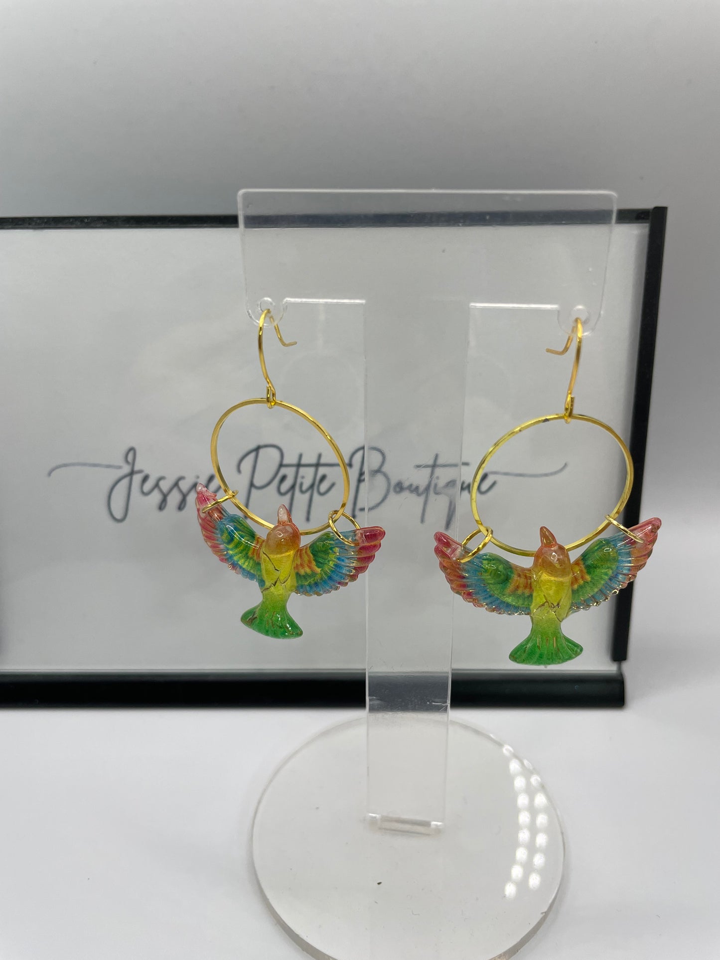Colorful bird-shaped earrings on a clear stand with a branded box in the background.