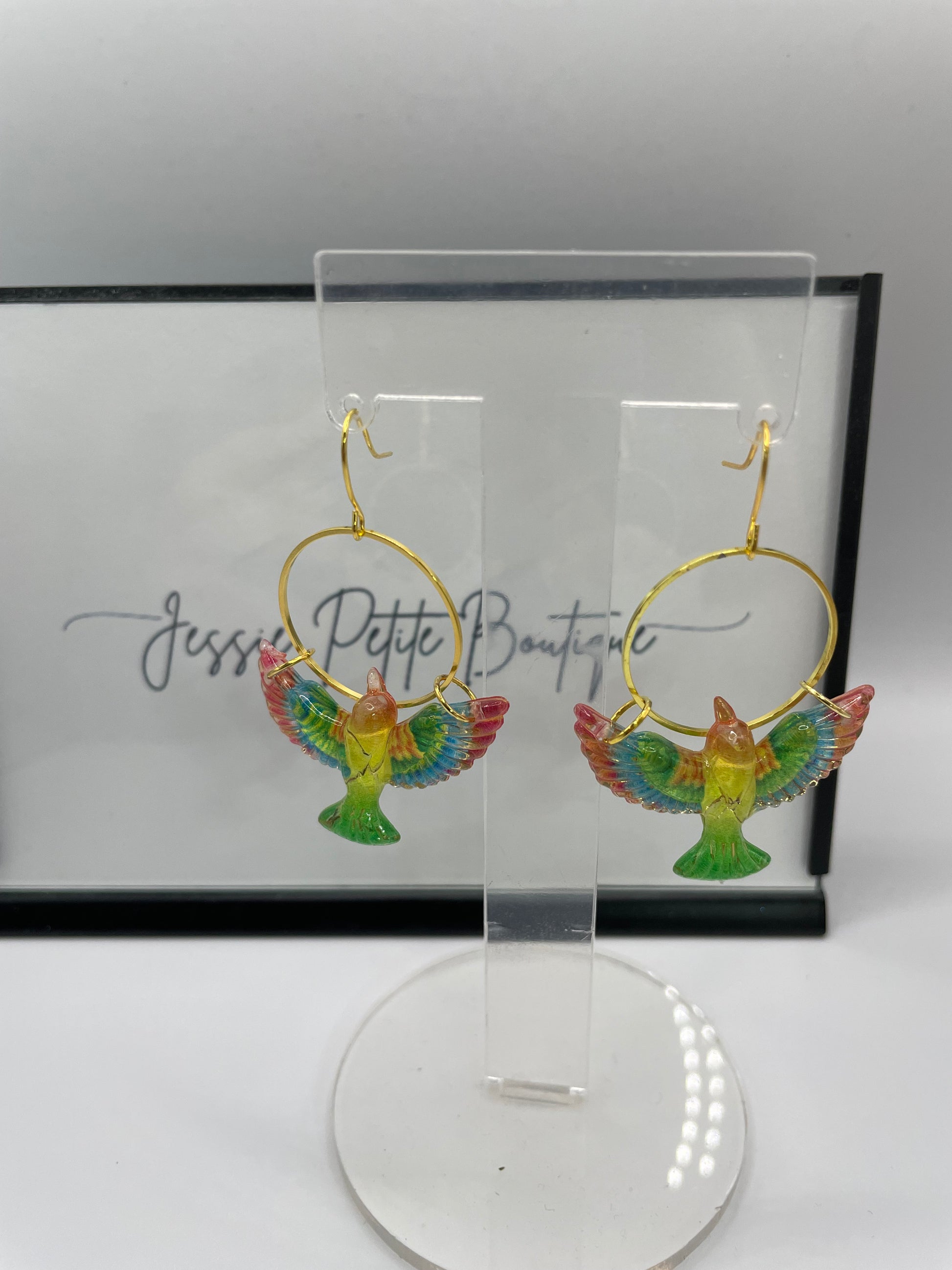 Colorful bird-shaped earrings on a clear stand with a branded box in the background.