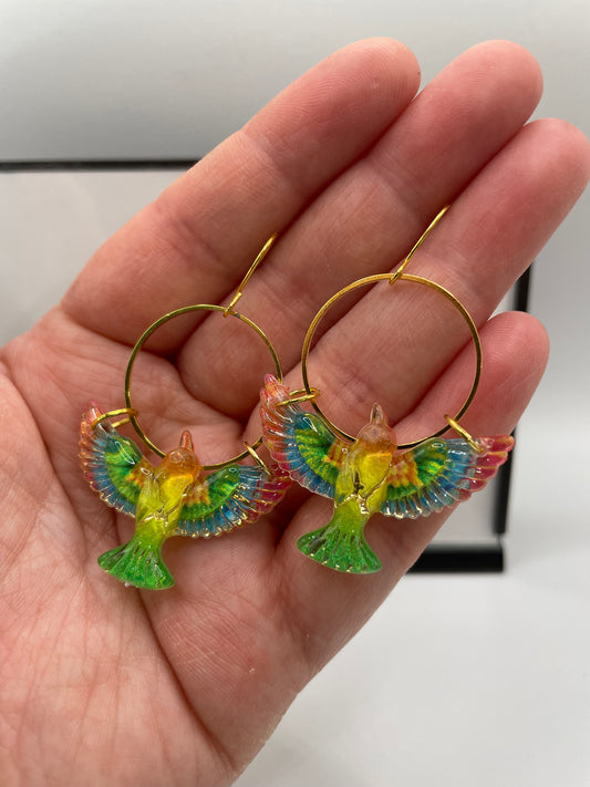 Hand holding colorful bird-shaped hoop earrings against a neutral background