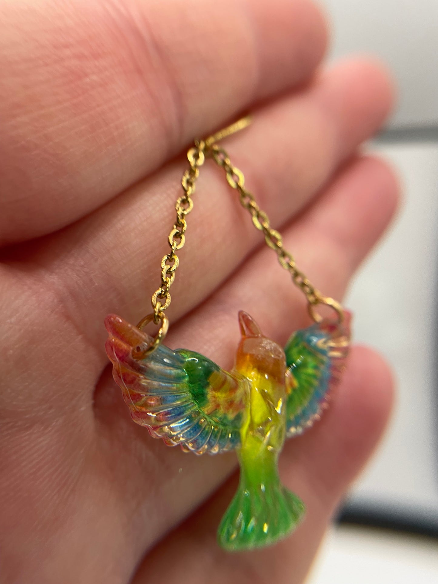 Colorful bird-shaped pendant on a gold chain held in a hand.