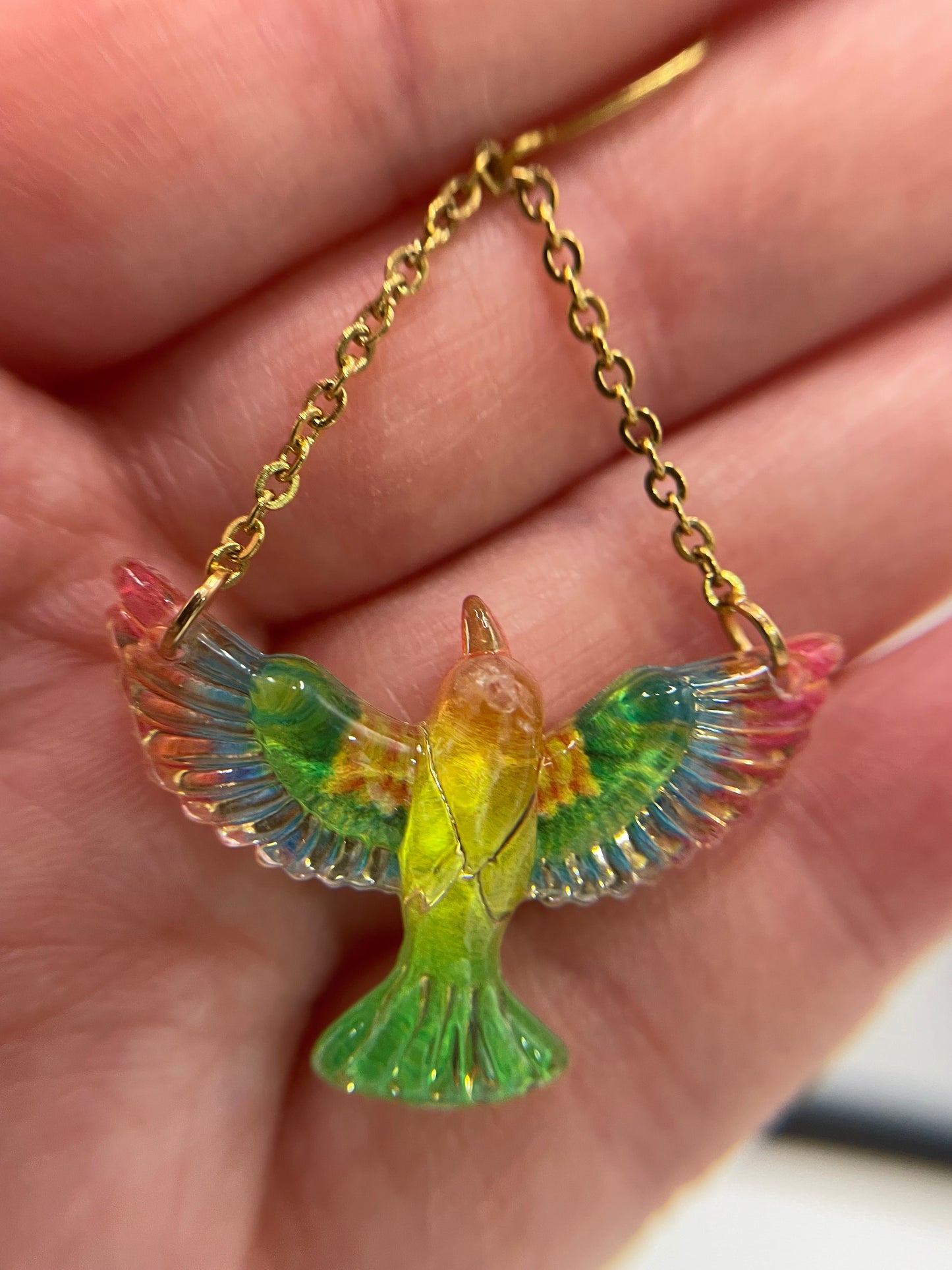 Colorful bird-shaped pendant on a gold chain held in a hand.