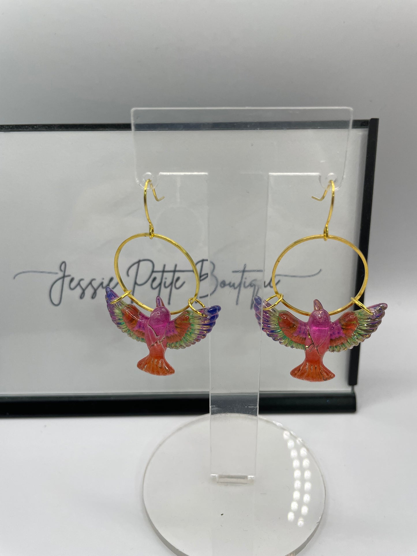Colorful bird-shaped earrings in a clear display case with a brand name on a white background.