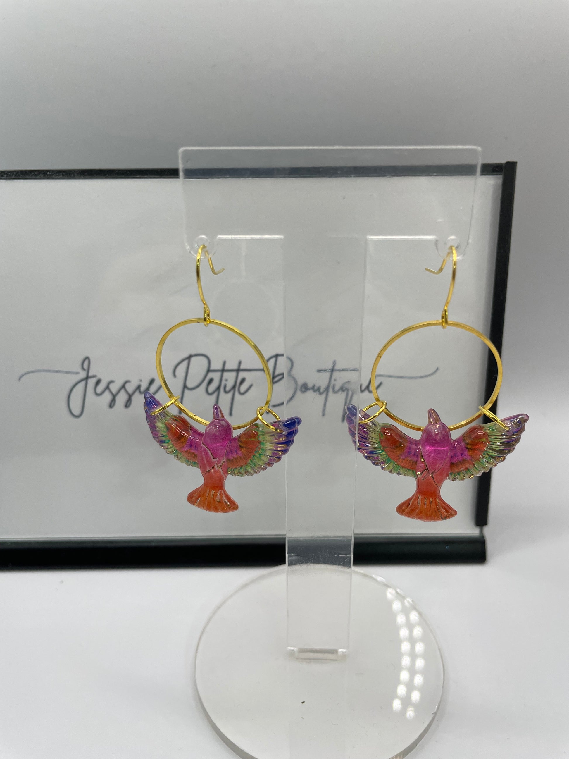 Colorful bird-shaped earrings in a clear display case with a brand name on a white background.
