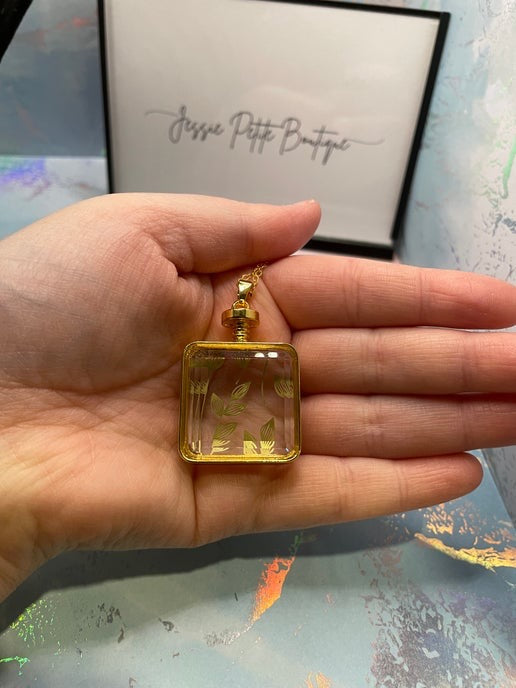 Hand holding a gold necklace with a clear pendant featuring leaves, against a blurred background.