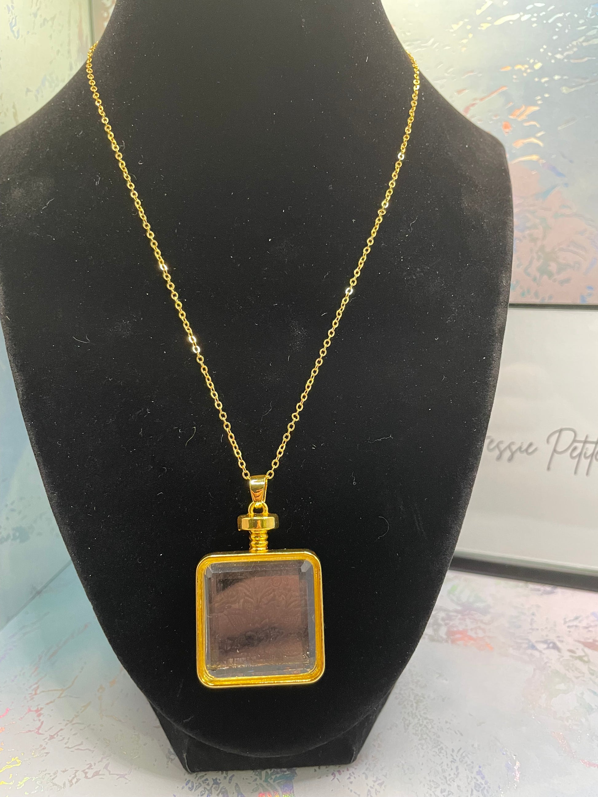 Gold necklace with a square pendant on a black stand.