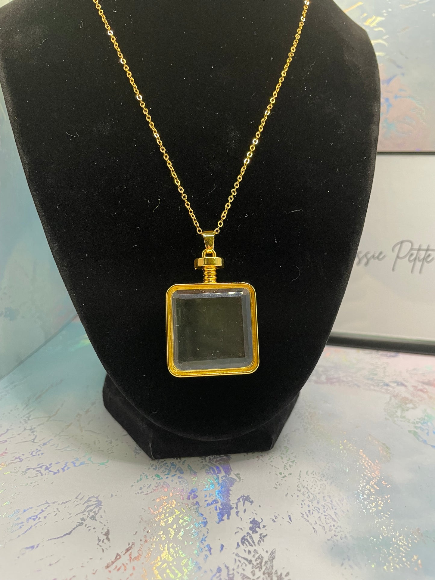 Gold necklace with a square pendant on a black stand.