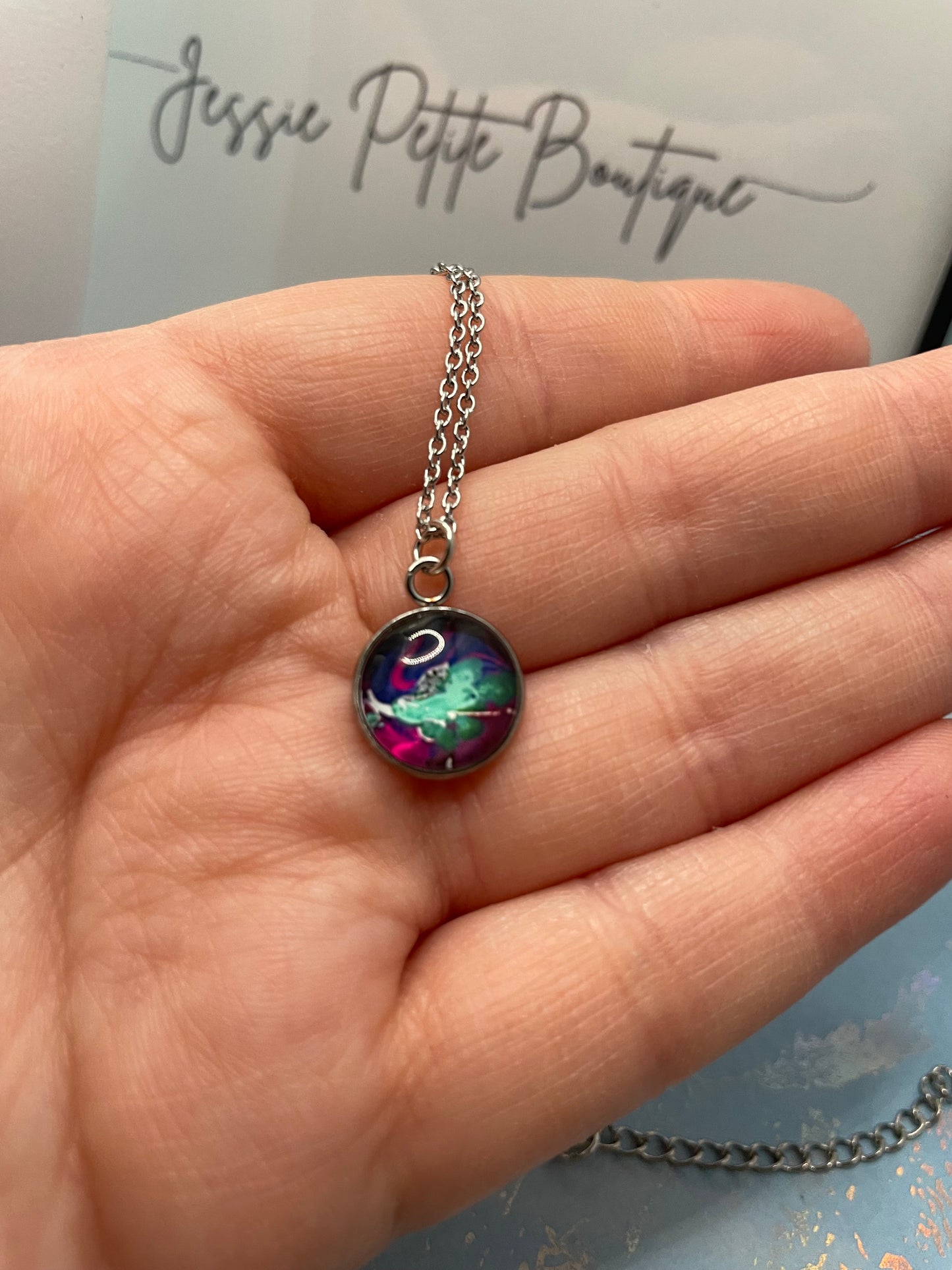 Necklace with a colorful pendant held in a hand, with 'Jessie Petite Boutique' in the background.