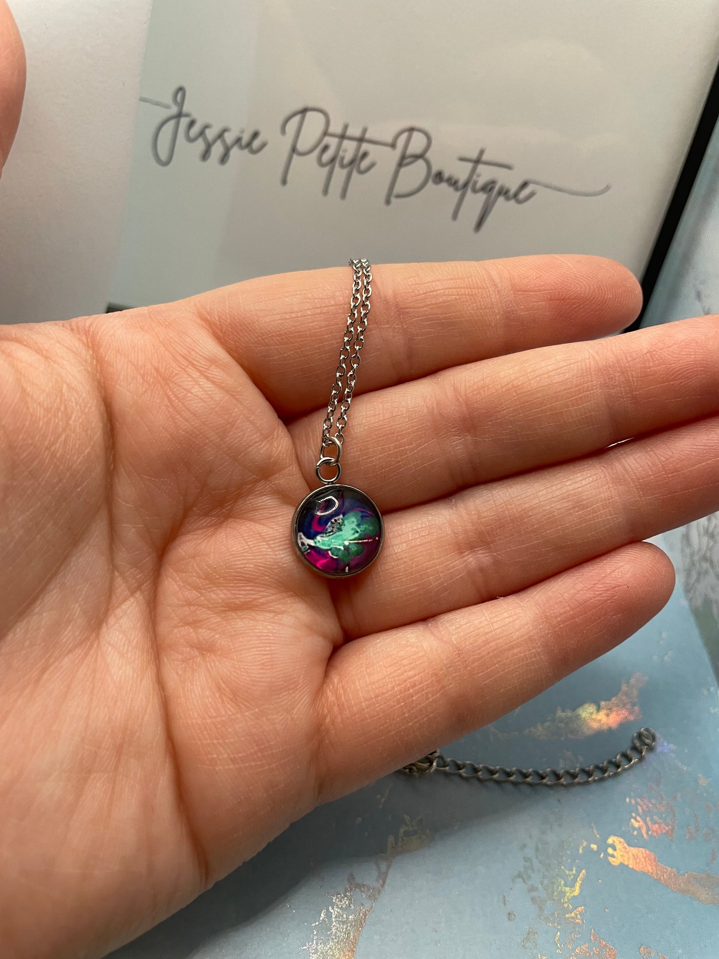 Hand holding a necklace with a colorful pendant, with 'Jessie Petite Boutique' in the background.