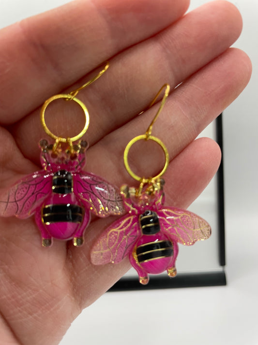 Pink bee-shaped earrings with gold accents held in a hand against a white background