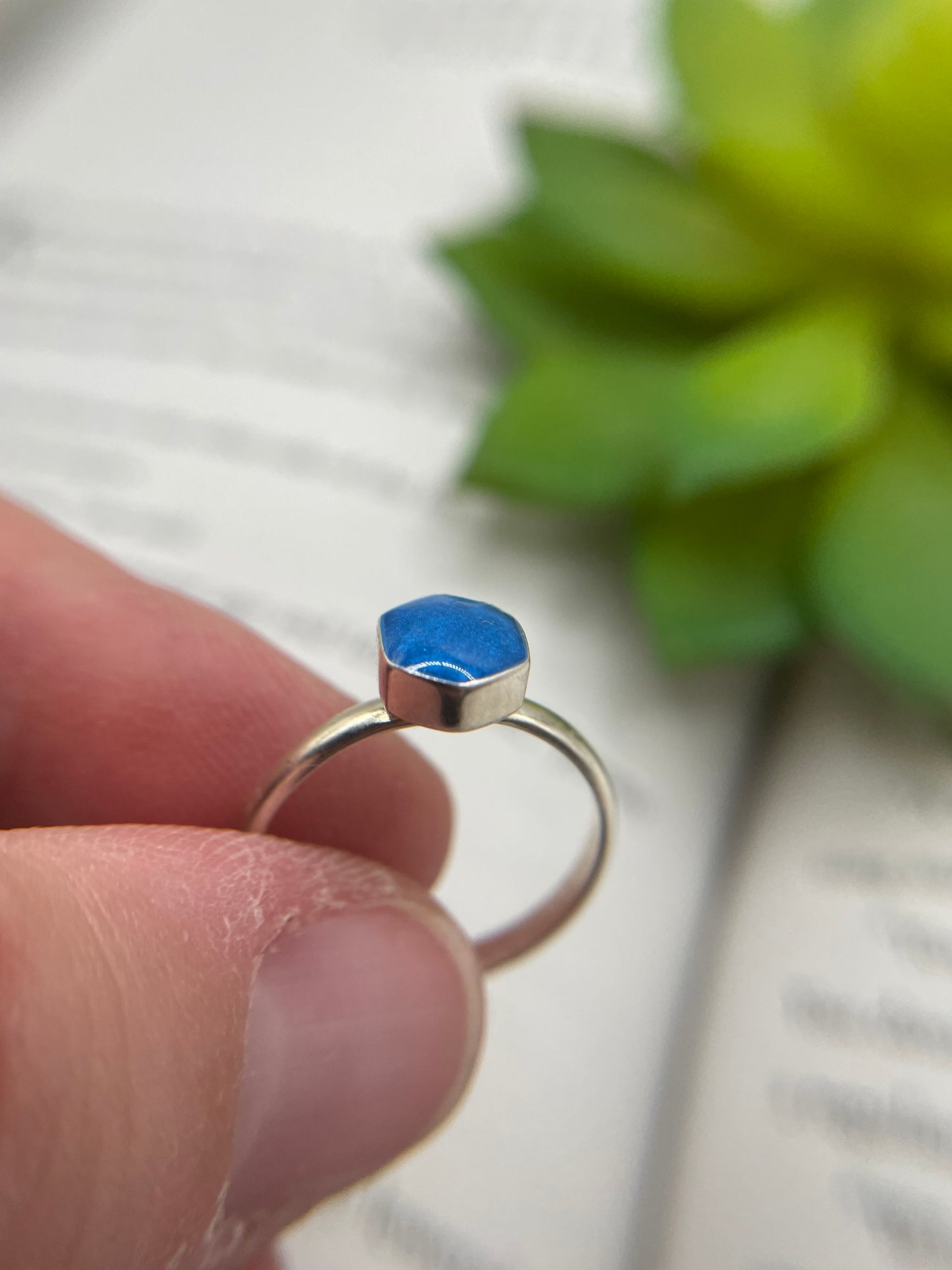 Silver ring with a blue gemstone held between fingers against a blurred green plant background
