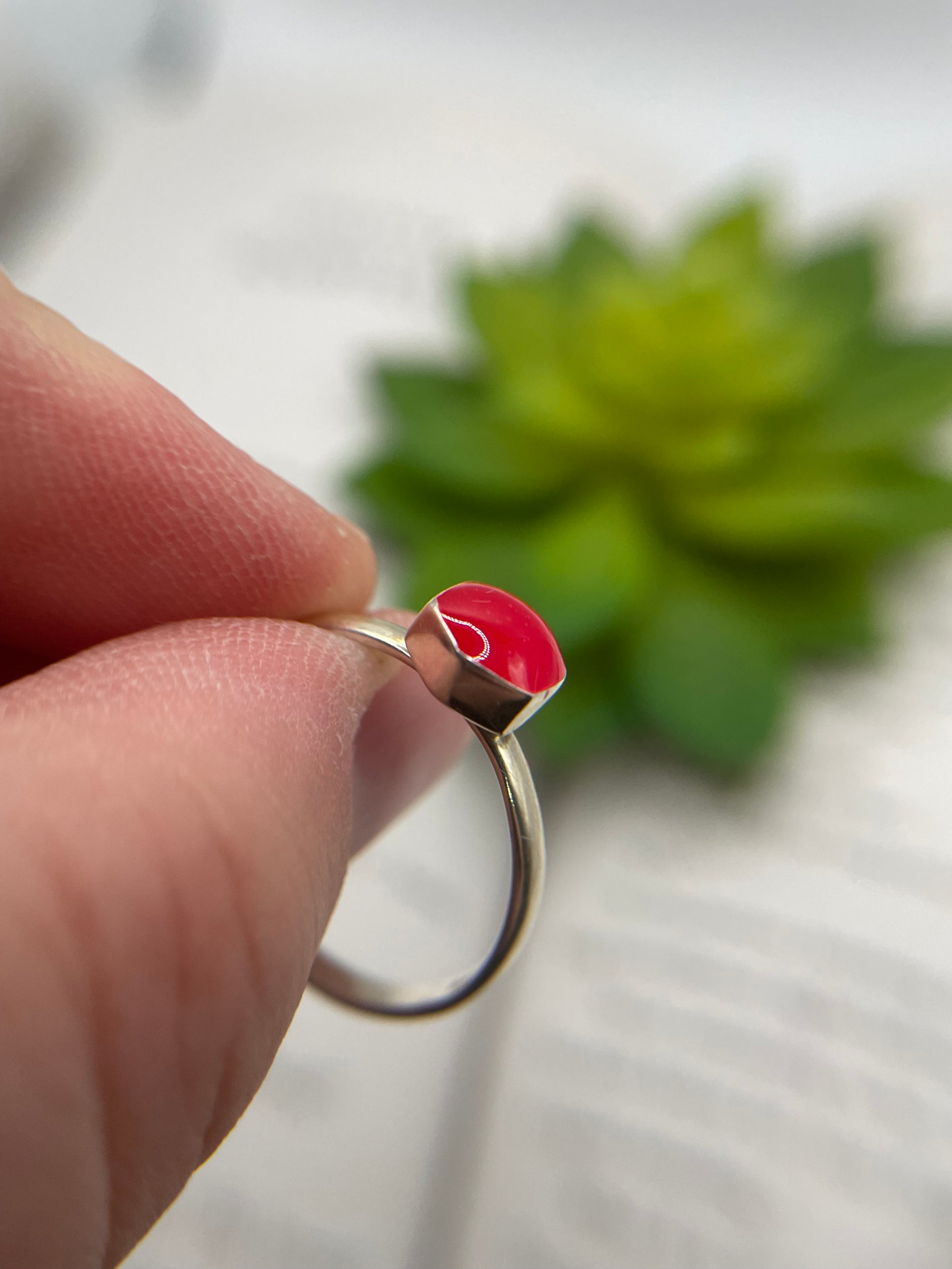Close-up of a hand holding a silver ring with a red stone against a blurred green plant background.