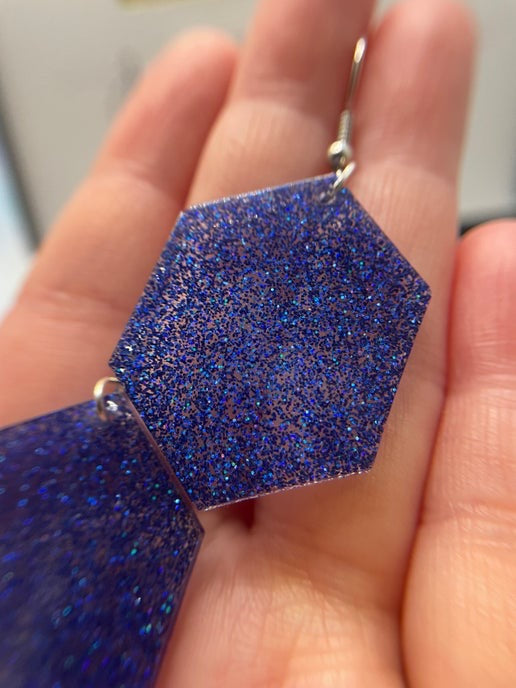 Blue glittery earrings held in a hand against a neutral background